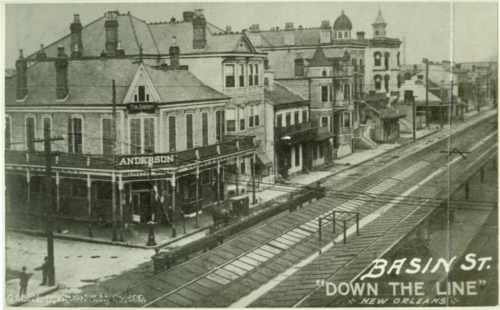 Black and white vintage photo of Basin Street in New Orleans. The street is lined with historic buildings, including Anderson and The Annex. Trolley tracks run along the road, and pedestrians walk on the sidewalks.