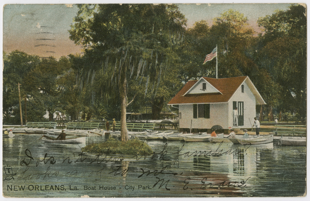 A vintage postcard depicts a boathouse on a tranquil lake in City Park, New Orleans. Several rowboats float near the boathouse, which is surrounded by lush trees. An American flag flies atop the boathouse on a sunny day. Handwritten text overlays the image.