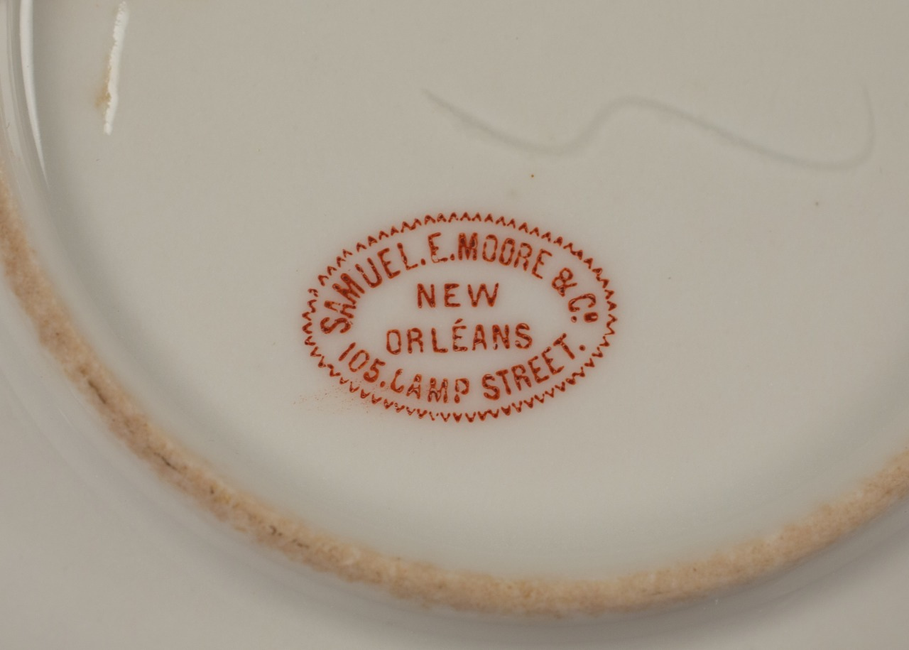 Close-up of a stamped marking on ceramic, reading SAMUEL E. MOORE & CO. NEW ORLEANS 105 LAMP STREET in red text inside an oval shape.