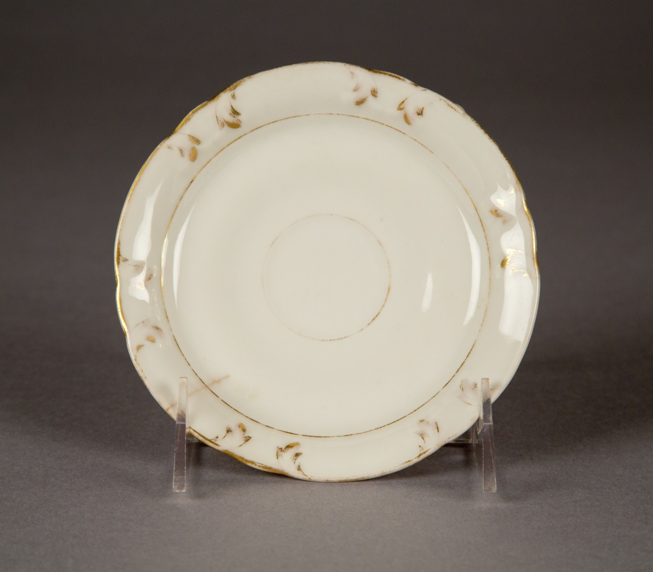 A white porcelain saucer with a subtle floral pattern along the wavy rim, accented by a thin gold trim. The saucer is displayed on a transparent stand against a neutral background.