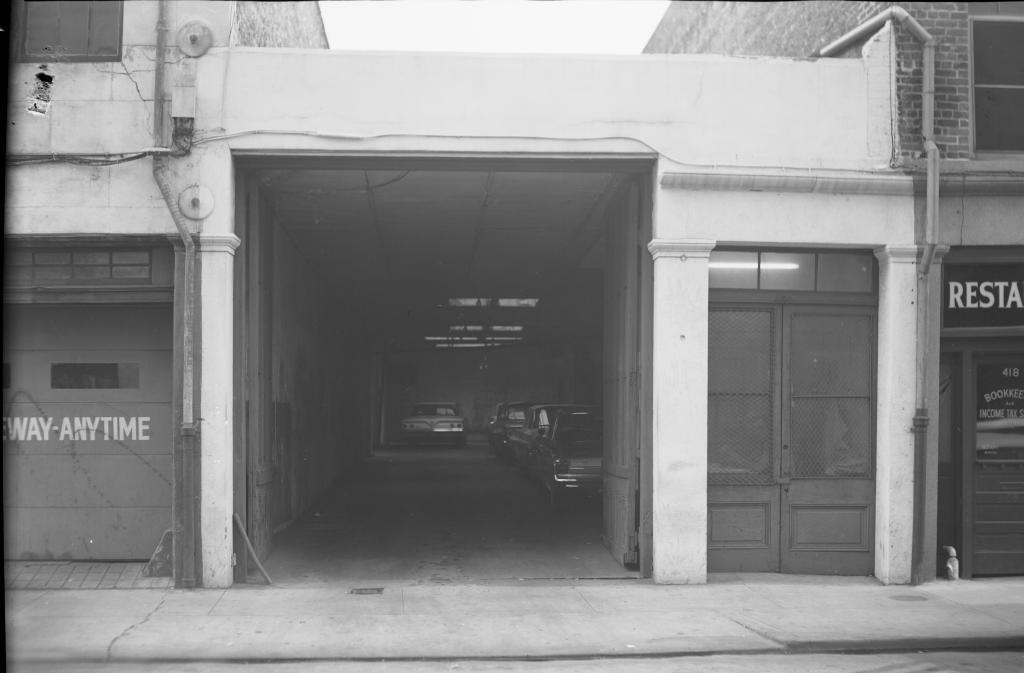 A black and white photo of a narrow garage entrance with cars parked inside. The building has a partially open door on the right, and a sign on the left reads ANYTIME. Its surrounded by brick walls and an industrial facade.
