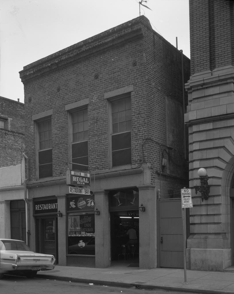 Black and white photo of a brick building housing The Regal restaurant and bar. The signage includes a menu board in the window. A car is parked in front on the street, and a No Parking sign is visible.