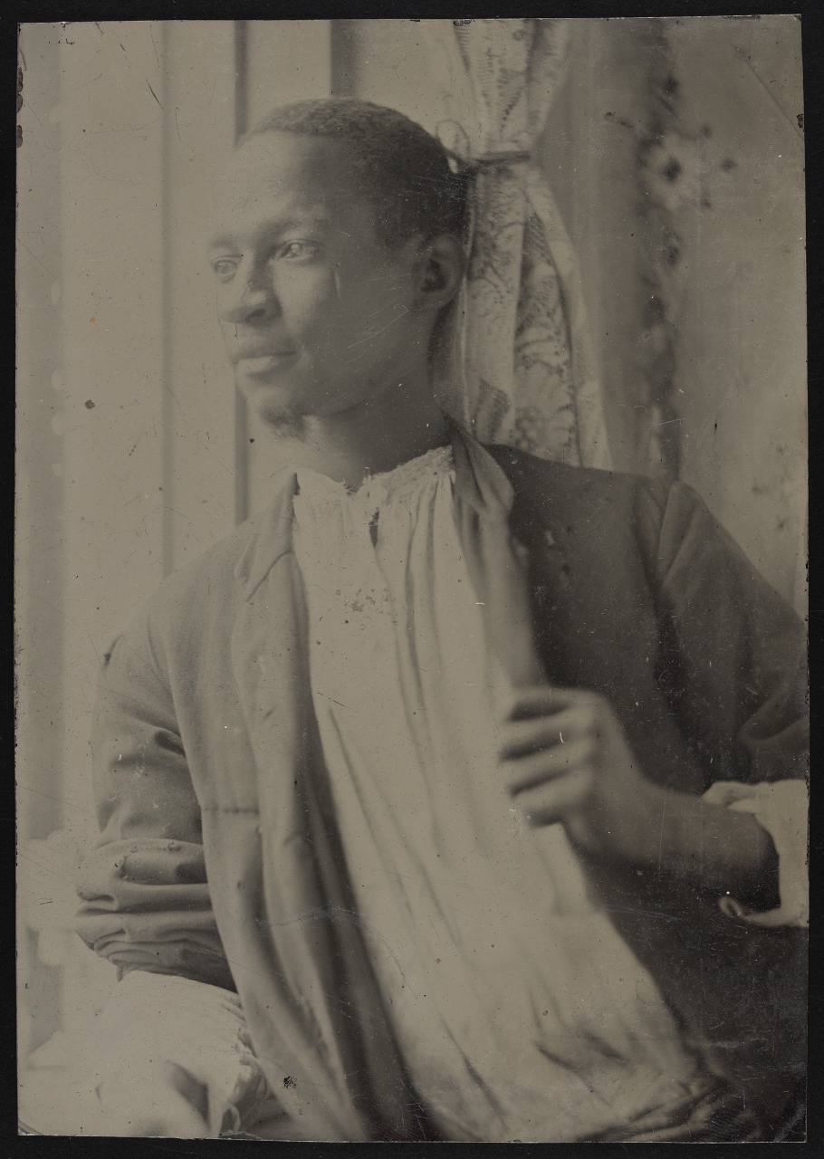 A black and white photo of a man wearing a loose, light-colored shirt and dark jacket, looking thoughtfully out a window. The background includes partially drawn curtains.