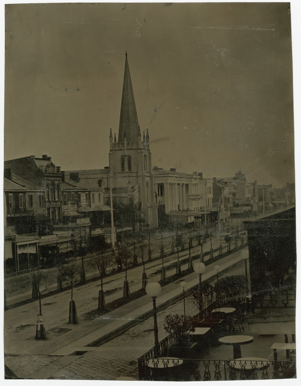 A sepia-toned vintage photograph of a city street featuring a prominent church with a tall spire. The street is lined with historic buildings and streetlamps, with a small outdoor seating area visible in the foreground.