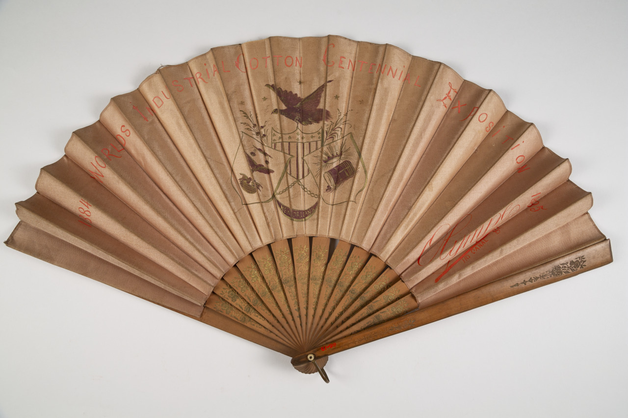 A decorative antique hand fan featuring a coat of arms with an eagle and shield in the center. The fan is inscribed with International Cotton Centennial, and has a light brown, textured finish with subtle historical motifs.