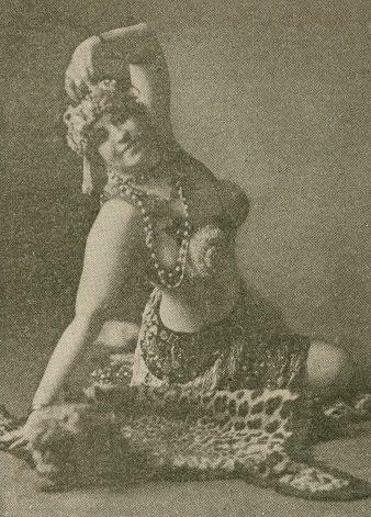 A vintage photo of a woman in a decorative costume with elaborate headwear and jewelry. She is striking a graceful pose while seated on a leopard skin rug, evoking an exotic and artistic ambiance.