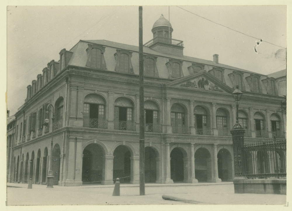 A black and white photograph of a historic, two-story building with arched windows and a dome on the roof. The architecture features ornate details and a staircase leading to the entrance, surrounded by street lamps and a fence.