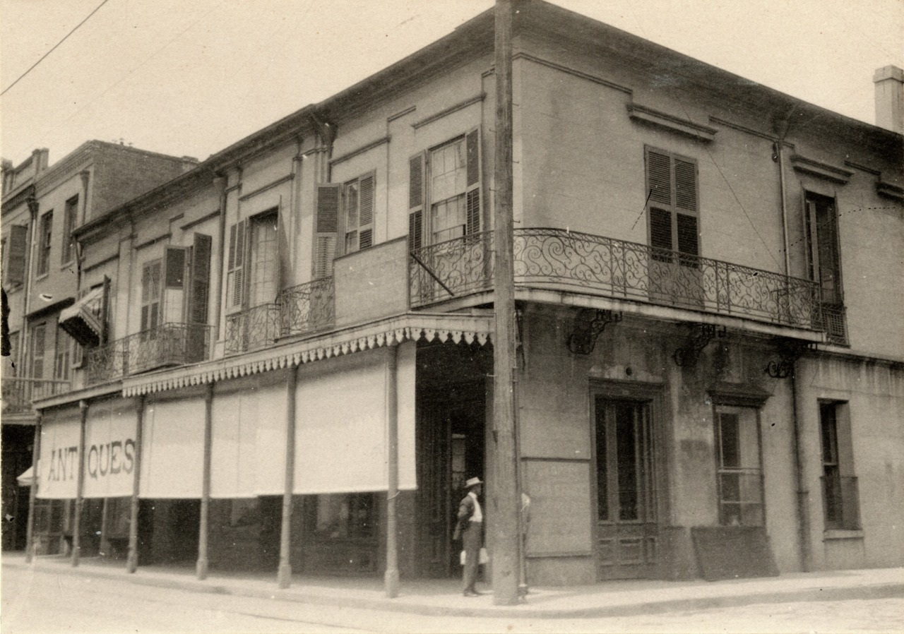 A vintage photograph of a two-story building with a wraparound balcony featuring ornate wrought iron railings. The ground floor has large windows, an awning with the word ANTIQUES, and a person standing nearby on the corner.