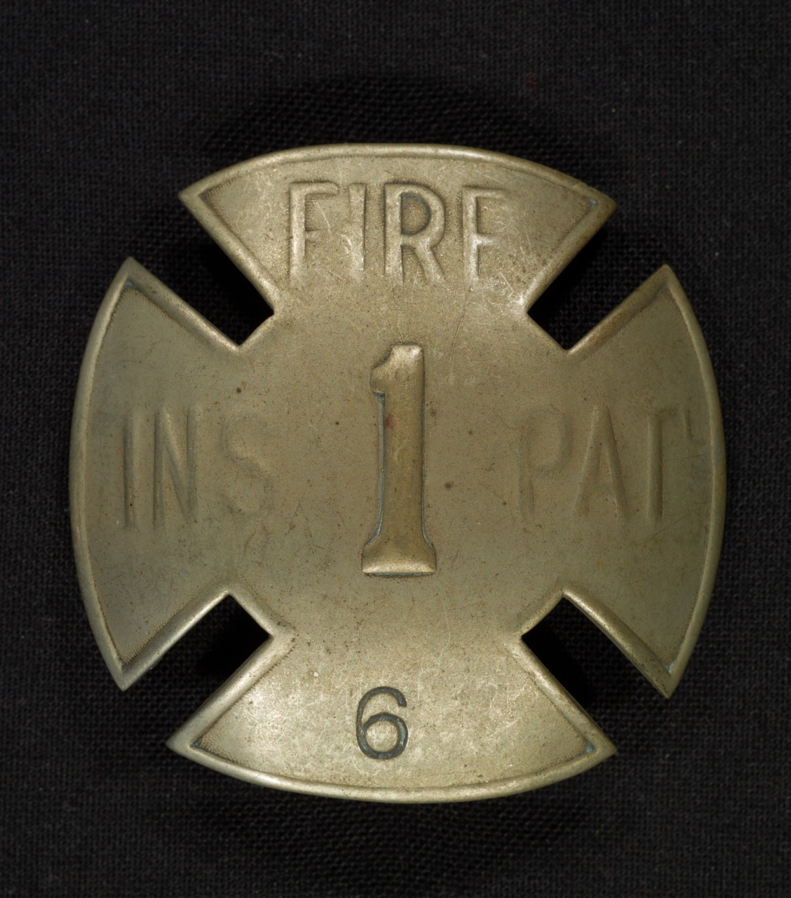 A brass Maltese cross-shaped badge with the word FIRE at the top, a large number 1 in the center, and a number 6 at the bottom. The background is black.
