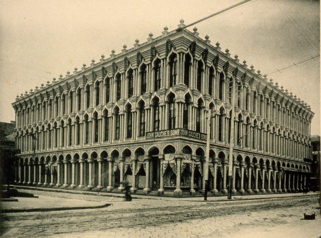 A historic, multi-story building on a cobblestone street corner, featuring long rows of ornate arched windows. Signage reads John Bouch & Sons. The architecture is detailed and symmetrical.