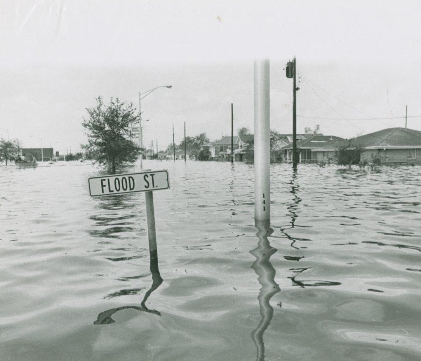 A street submerged in floodwater, with a partially submerged Flood St. sign in the foreground. Houses and trees are visible in the background under a cloudy sky.