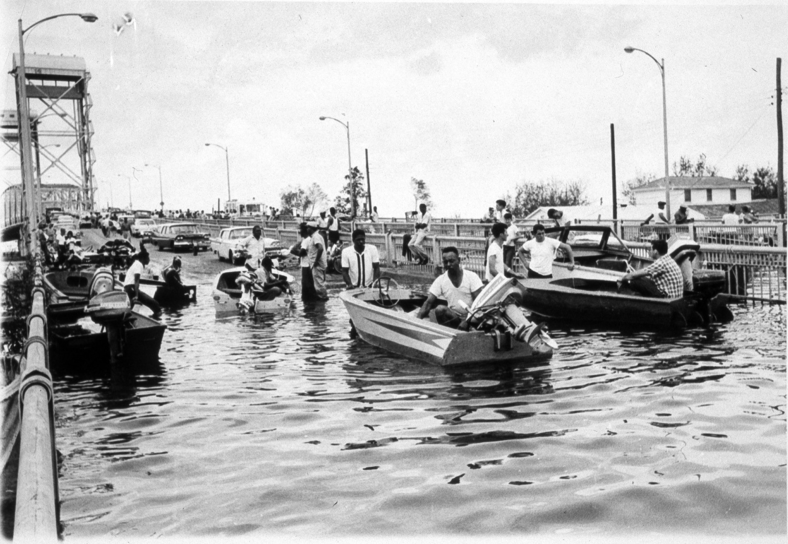 People in small boats navigate a flooded street, with cars partially submerged. The scene includes several onlookers and structures visible in the background. The sky is overcast, indicating a likely aftermath of heavy rain or a storm.