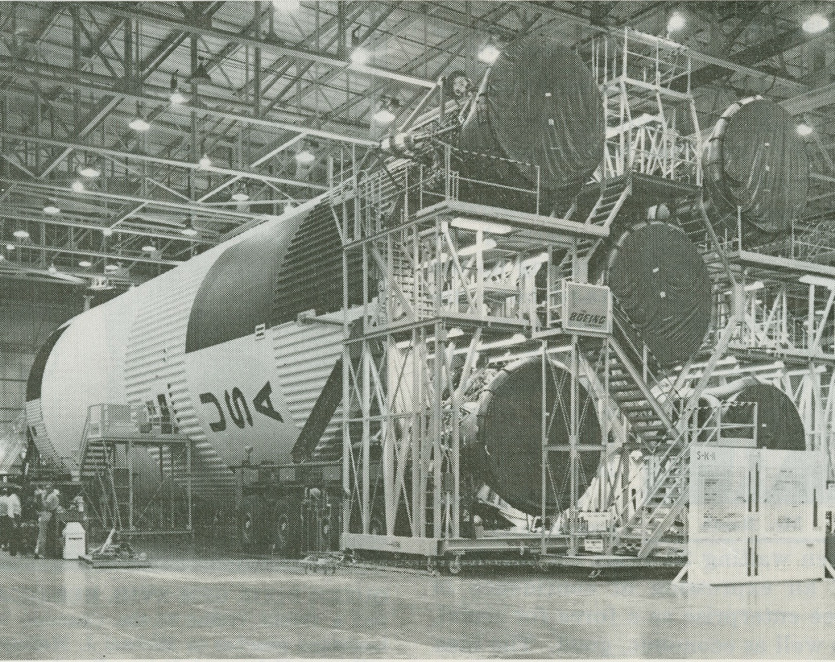 A black and white photo of a large spacecraft assembly area, featuring sections of a Saturn V rocket. Multiple scaffolds and platforms surround the rocket, with workers positioned around the structure in a spacious indoor facility.