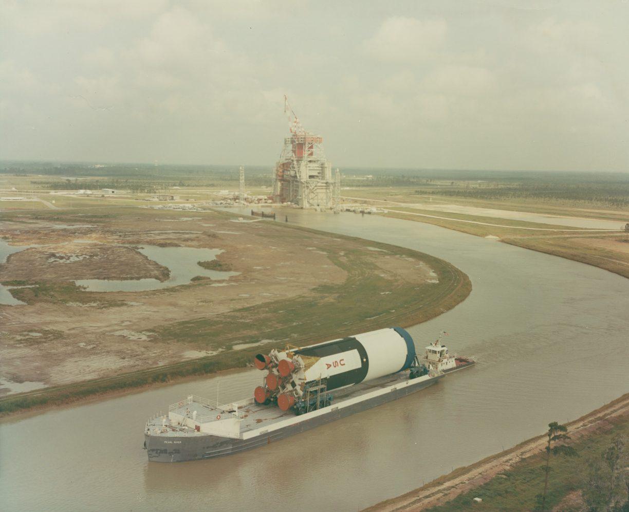 A barge carrying a large rocket booster sails along a wide canal, bordered by grassy banks. In the background, there is a large industrial structure with cranes, under a partly cloudy sky.