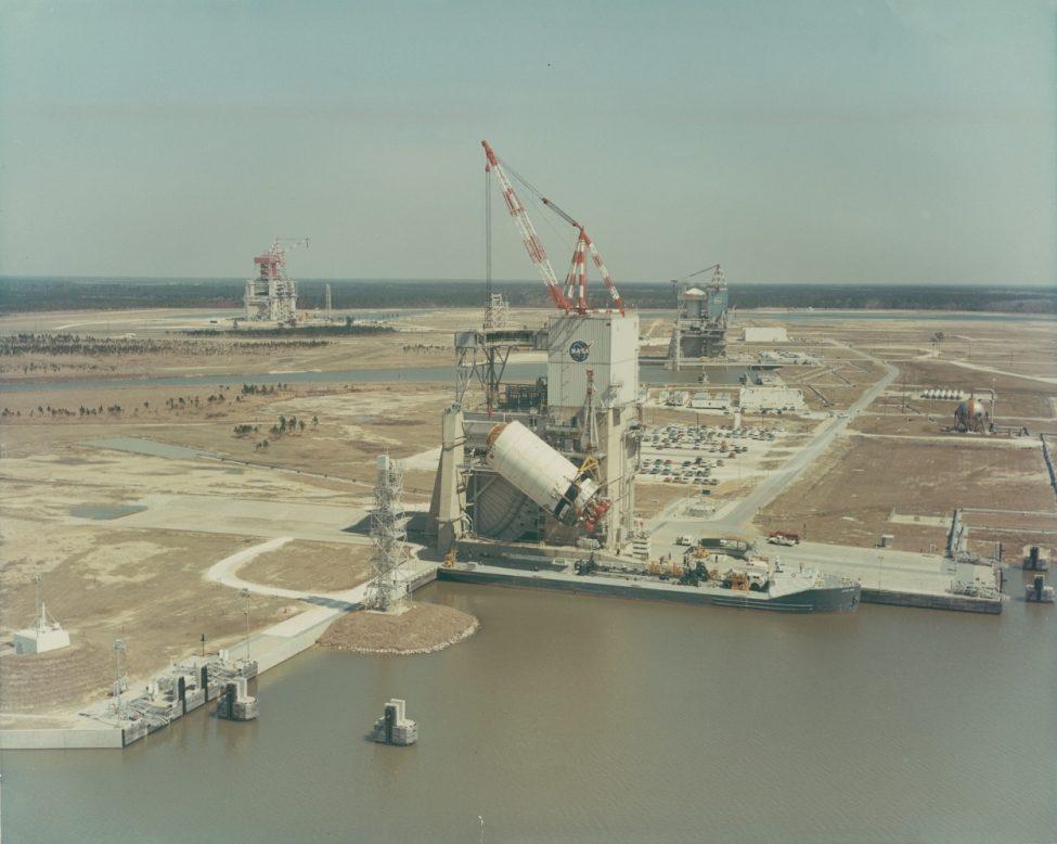 A large NASA rocket engine test stand is situated near a waterway. The area is surrounded by flat land with scattered buildings and cranes. The sky is clear, and the scene captures the expansive and industrial nature of the space facility.