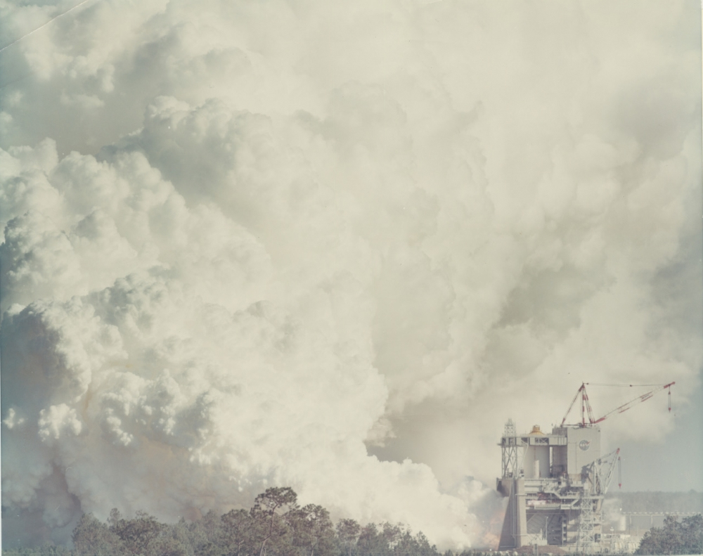 A large cloud of smoke billows from a launch pad area amid trees and industrial structures, set against a pale, cloudy sky. Some cranes and machinery are visible near the launch site.