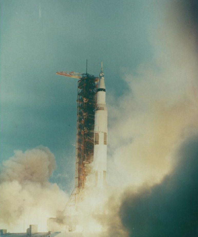 A large rocket is launching into the sky, surrounded by thick clouds of smoke and flames at its base. The background is a clear blue sky with a visible structure supporting the rocket on the left side.