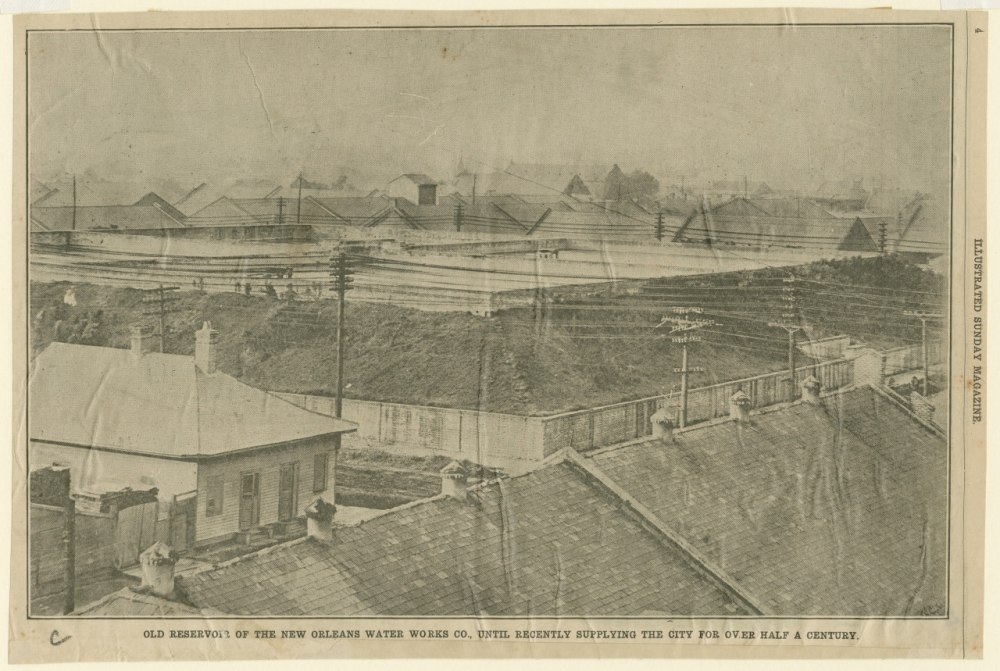 A historical photograph shows the old reservoir of the New Orleans Water Works Company. Multiple rooftops and telegraph poles are visible in the foreground, with the reservoir and surrounding buildings in the background under a cloudy sky.