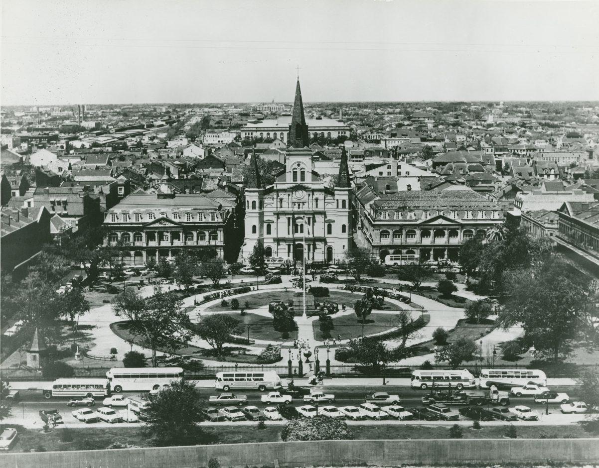 A vintage aerial view of Jackson Square in New Orleans, featuring St. Louis Cathedral in the center, surrounded by lush greenery and historic buildings. Several buses and cars are parked along the street in the foreground.