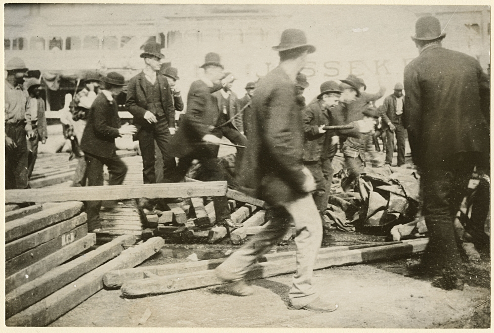 A historical black and white photo of several men in bowler hats, suits, and coats gathered on a busy street, possibly involved in loading or unloading activities with wooden beams scattered around. Buildings and a partially visible sign are in the background.