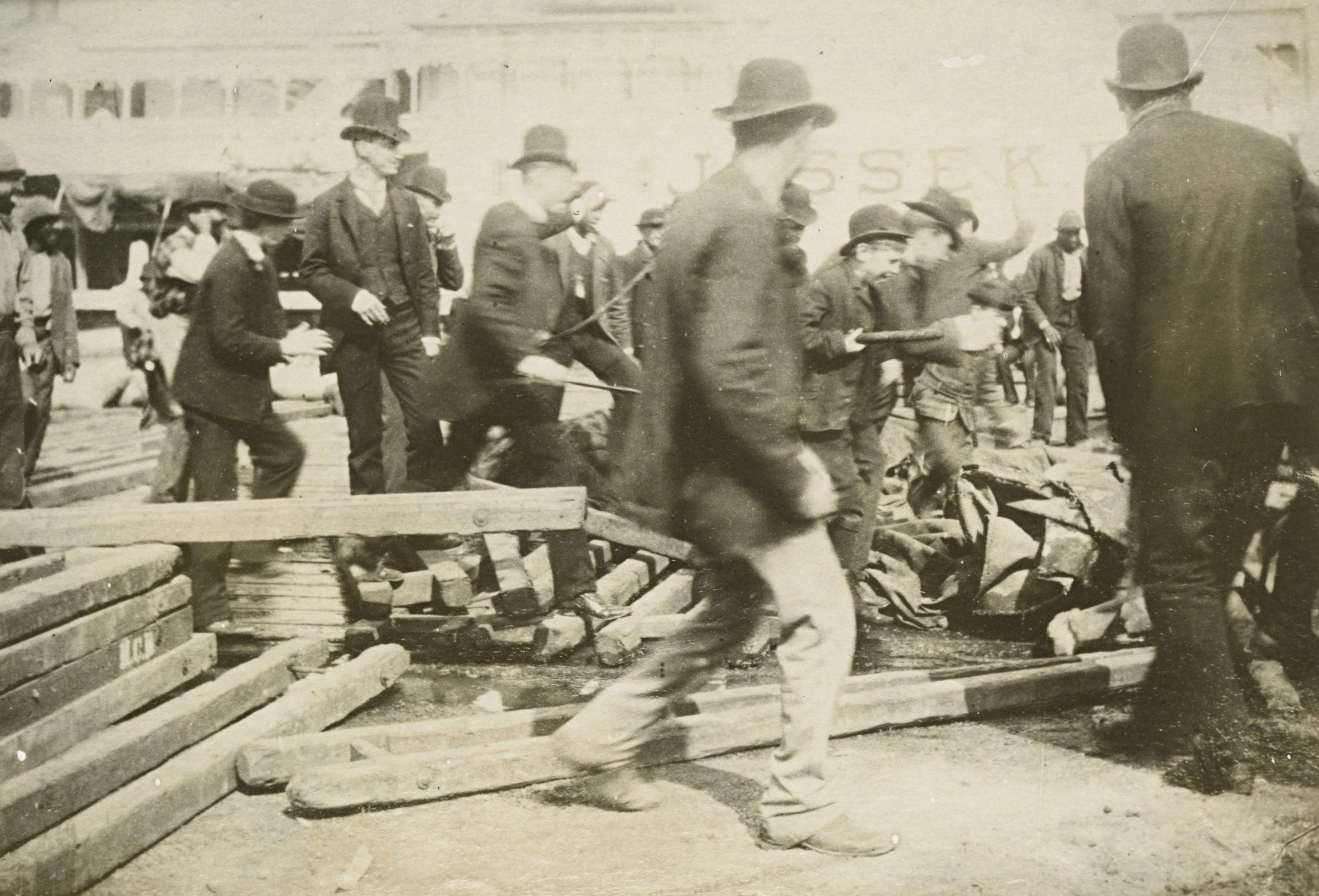 A historical black-and-white photo shows a group of men in bowler hats and suits, actively engaged in constructing or managing wooden beams on a busy dock. The scene conveys early 20th-century industrial activity.
