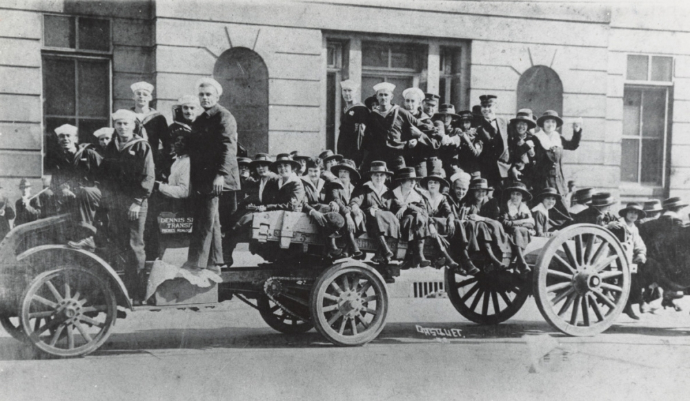 A vintage photograph shows a group of people, including men, women, and children, seated and standing on a large open vehicle with wheels. Theyre dressed in early 20th-century attire, with some waving. A building is visible in the background.