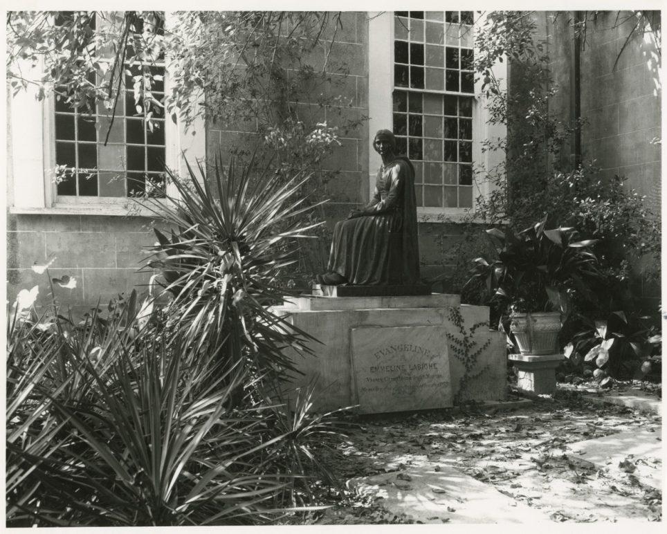 A historical black and white photograph of a seated statue in a garden. The statue is surrounded by lush plants and is situated in front of a stone building with large windows. Sunlight filters through the foliage, casting shadows on the scene.