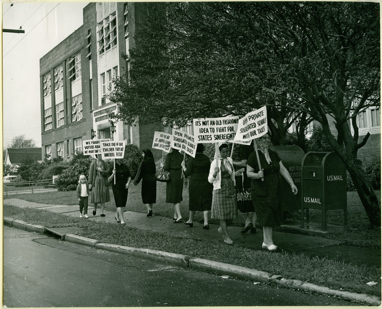 A group of women and children walk in a line on a sidewalk holding protest signs in front of a brick building. The signs address issues like states sovereignty and the federal school system. A large tree and U.S. Mail box are visible nearby.