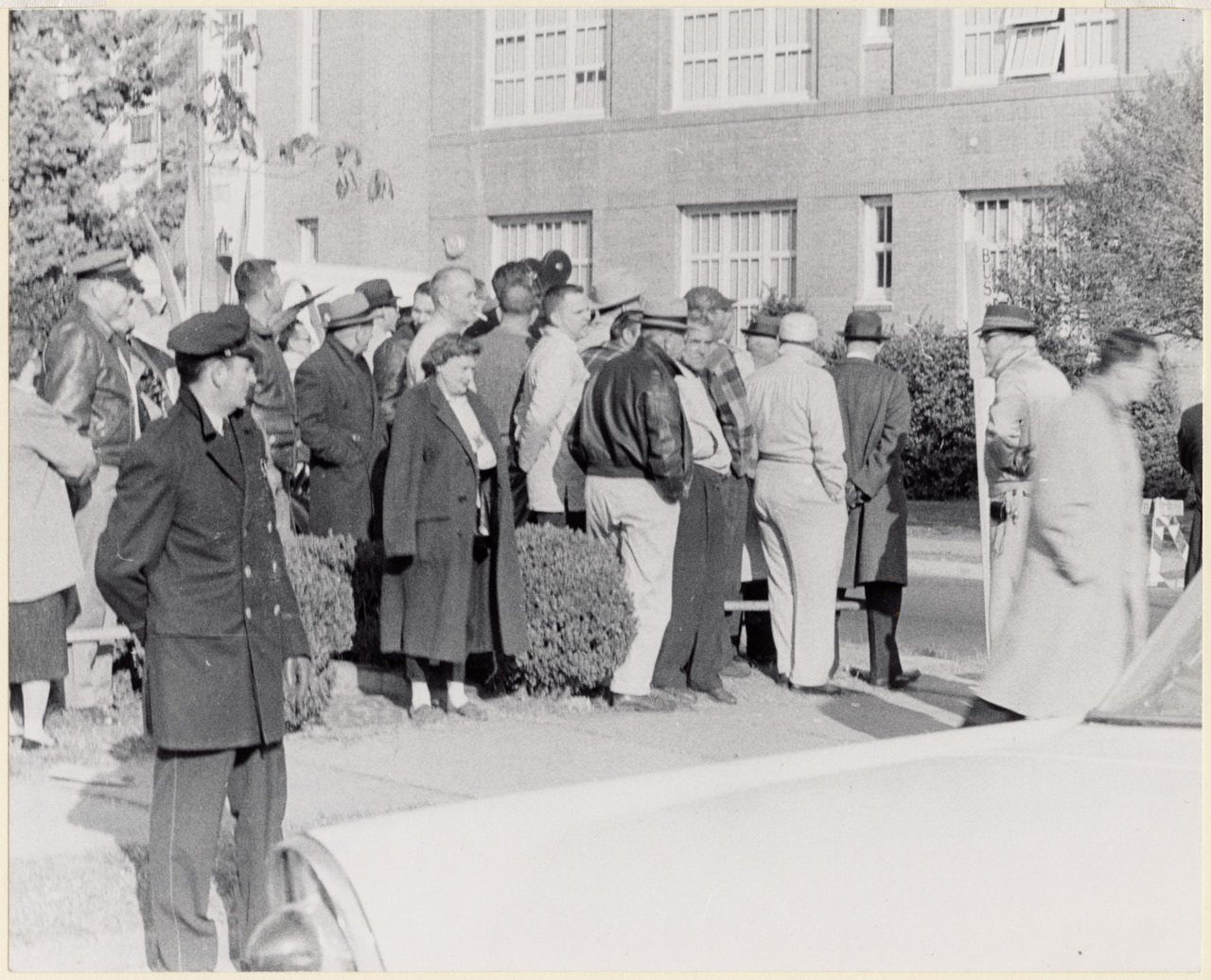 A group of people stands gathered on a sidewalk, facing a brick building. They are dressed in mid-20th century attire. A man in uniform stands in the foreground and another person walks by, blurred in motion.
