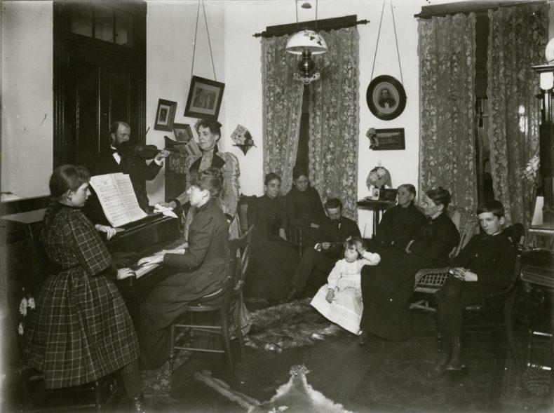 A black-and-white photo of a group of people gathered in a room. A woman plays the piano while others sit and listen. The setting includes ornate curtains, framed pictures, and a hanging light fixture. A child in white sits among the group.