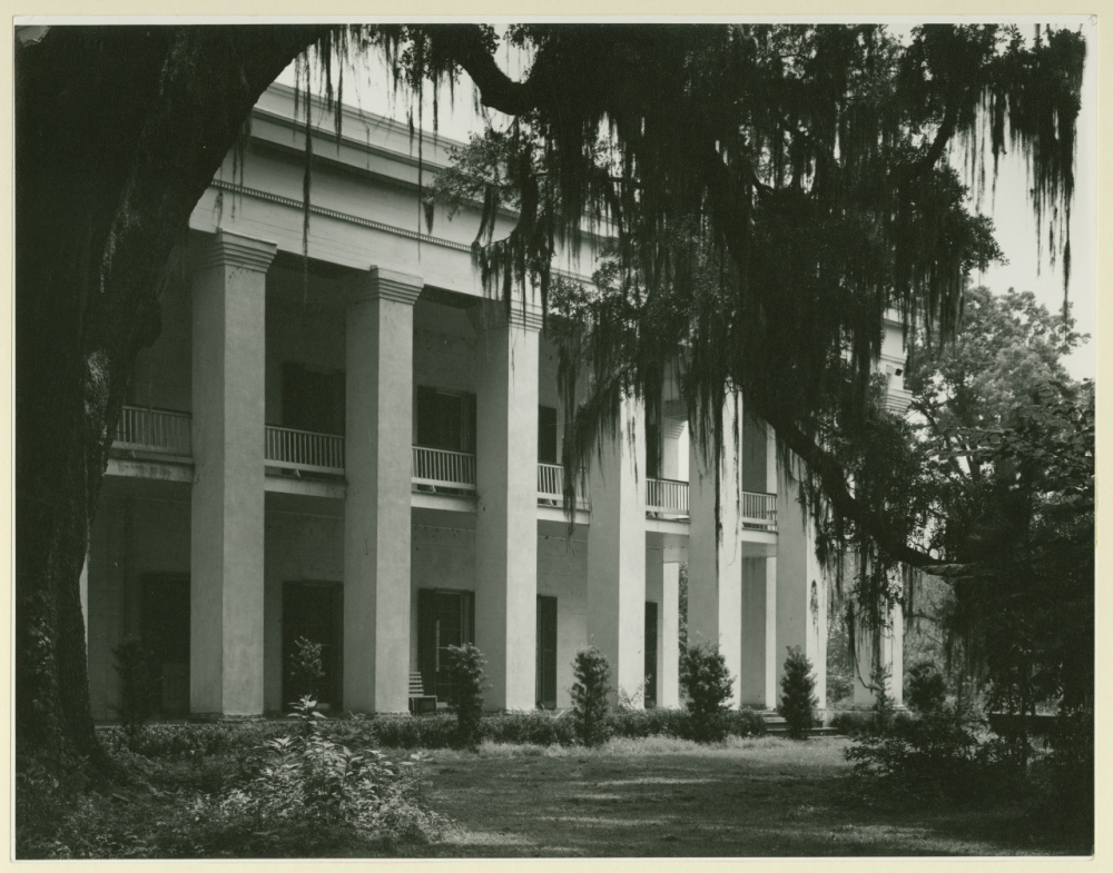 Black and white photo of a large, columned, neoclassical building surrounded by trees and shrubs. Spanish moss drapes from the branches, creating a historic and serene atmosphere.