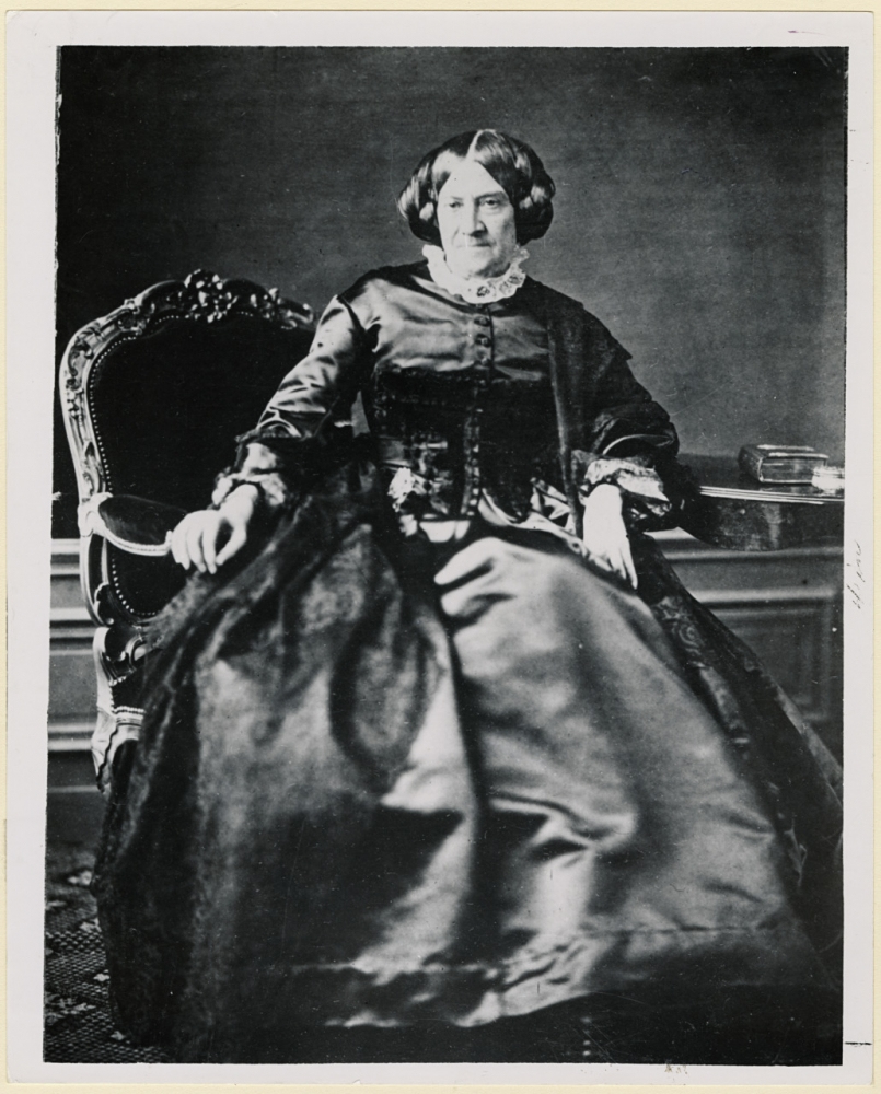 A sepia-toned portrait of a woman in formal 19th-century attire, seated on an ornate chair. She wears a long, dark dress with lace details and has a serious expression. A book rests on a table beside her.