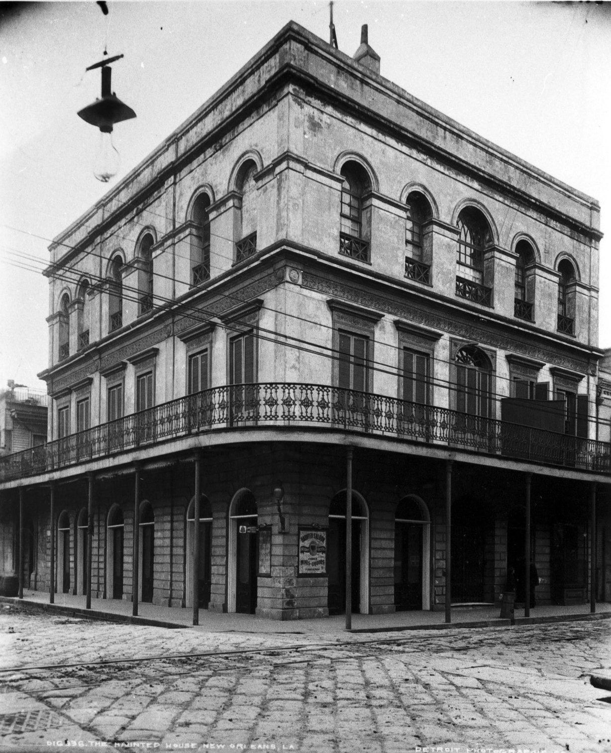 Black and white photo of a historic three-story building on a cobblestone street corner. It features arched windows and a wrought-iron balcony on the second floor. A streetlight hangs over the intersection.