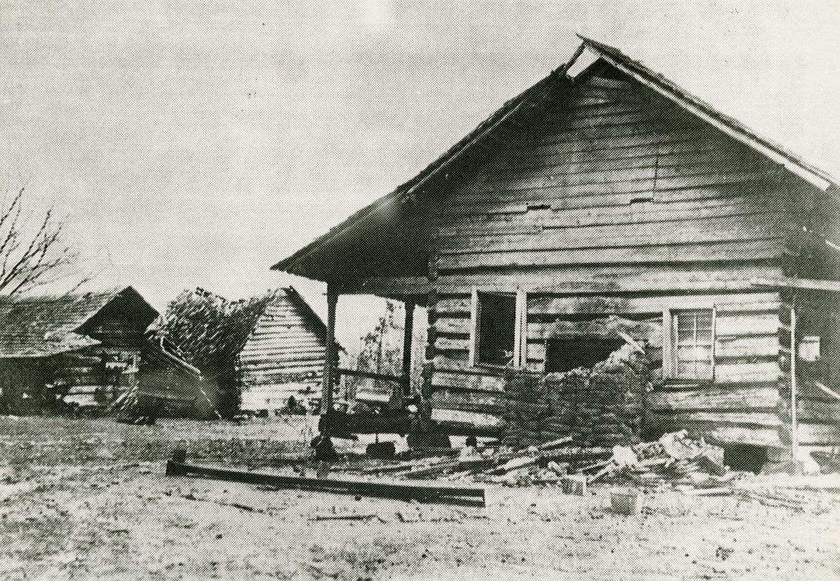 A vintage black-and-white photo of a wooden cabin with a partially collapsed wall. The building is surrounded by debris. A smaller structure is visible in the background on a barren landscape. Sky appears overcast.