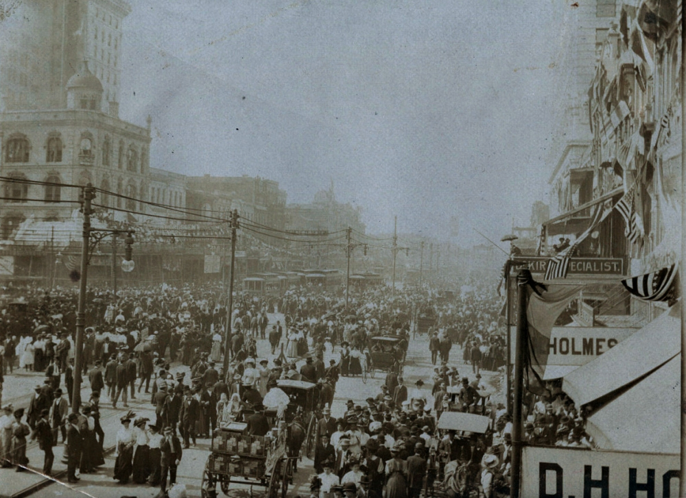 A historic photograph of a busy city street filled with people, horses, and carriages. Buildings line the street, with visible signs and banners. The atmosphere is bustling, suggesting an event or gathering in a bygone era.