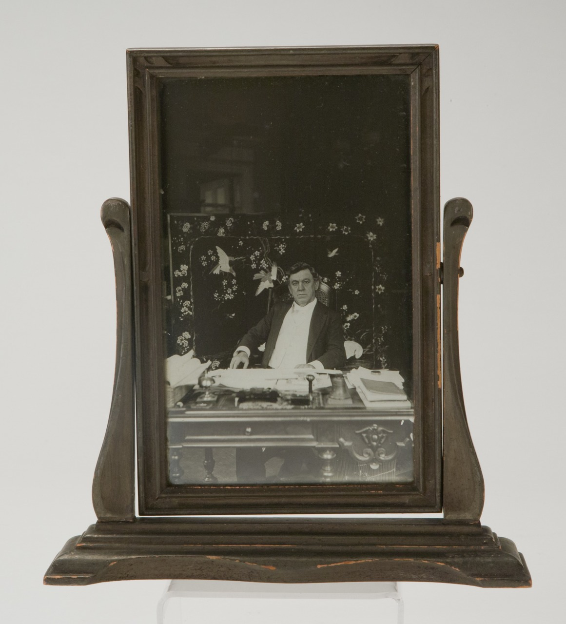 Vintage portrait of a person sitting at a desk with papers and a typewriter, framed in an ornate wooden stand. The background features a decorative screen with bird and floral patterns.