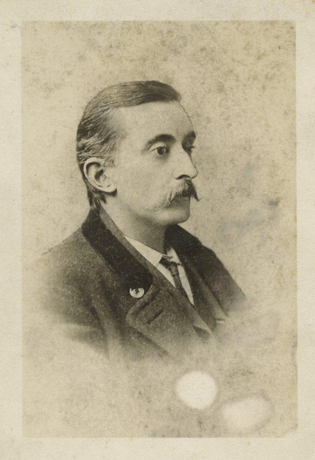 A sepia-toned vintage portrait of a man in profile, wearing a suit with a high-collared shirt and a tie. He has a mustache and short hair, and the background is lightly textured and faded.