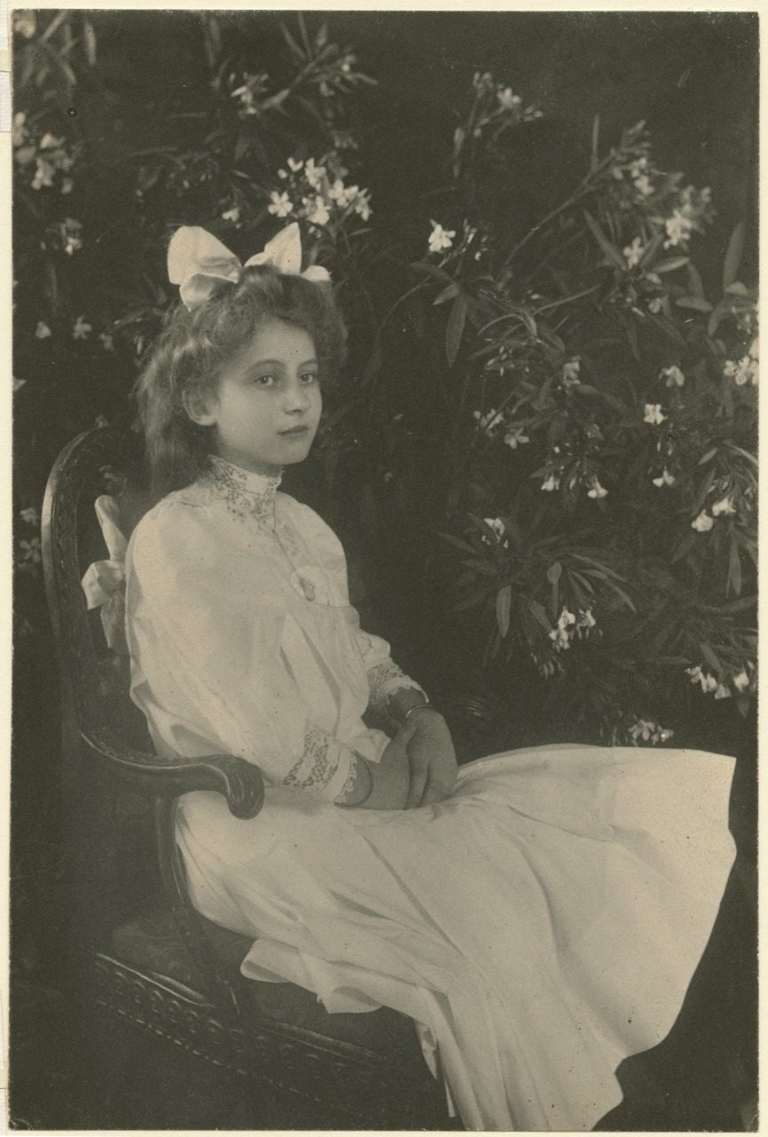 A young girl with long hair and a large bow sits in a wooden chair, wearing a white dress with lace details. She is surrounded by flowering plants, giving a serene, vintage atmosphere to the black-and-white photograph.