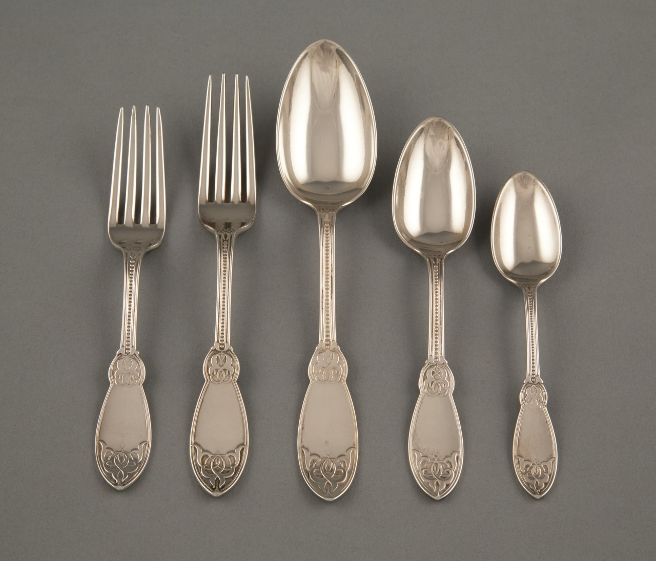 A set of five silver utensils arranged on a gray background, including two forks, a large spoon, a medium-sized spoon, and a small spoon, each with ornate detailing on the handles.