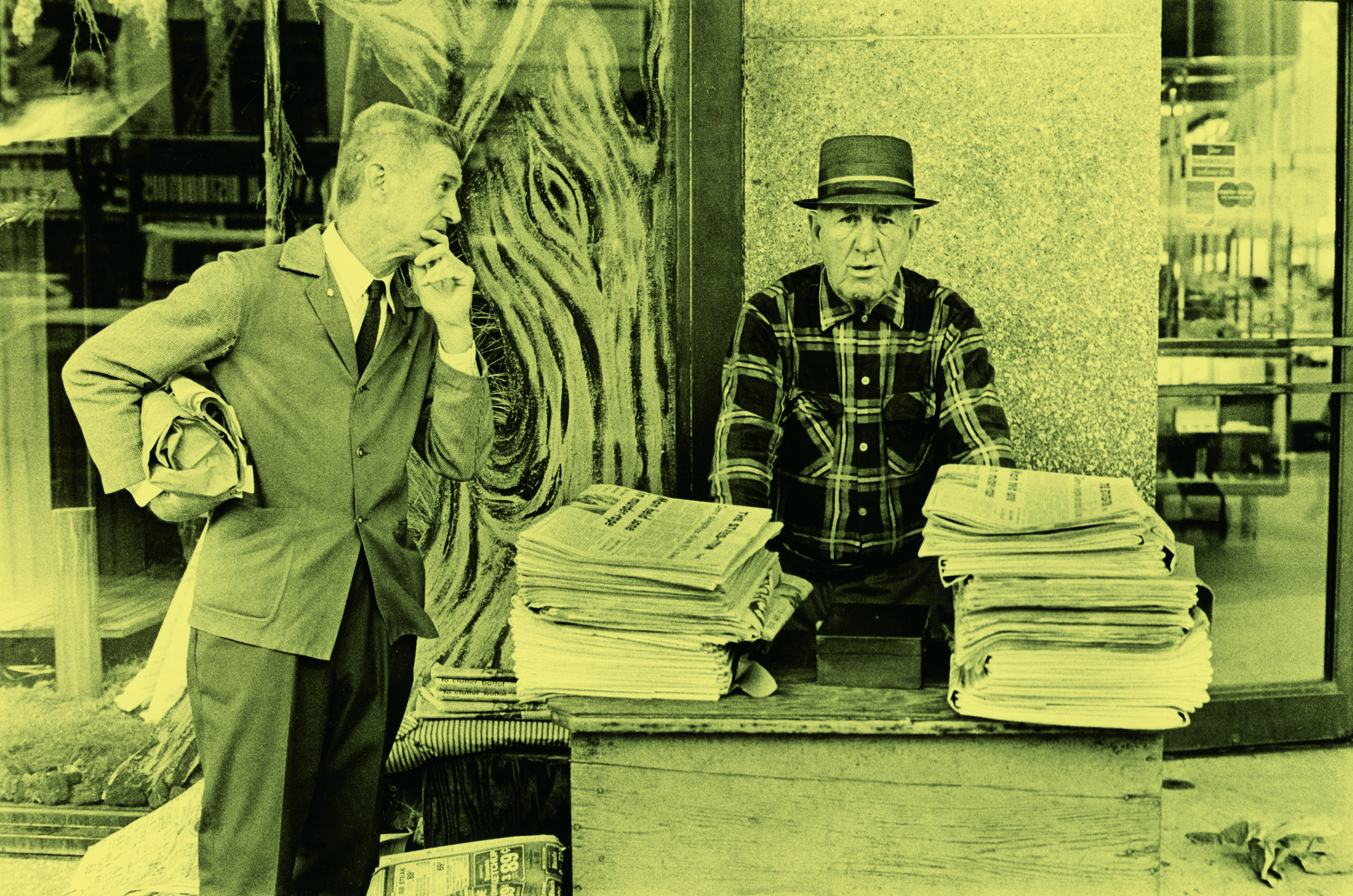 A man wearing a suit stands with newspapers under his arm, looking at a newsstand vendor. The vendor, in a plaid shirt and hat, stands behind a wooden counter piled with newspapers. The scene is set in front of a storefront window.