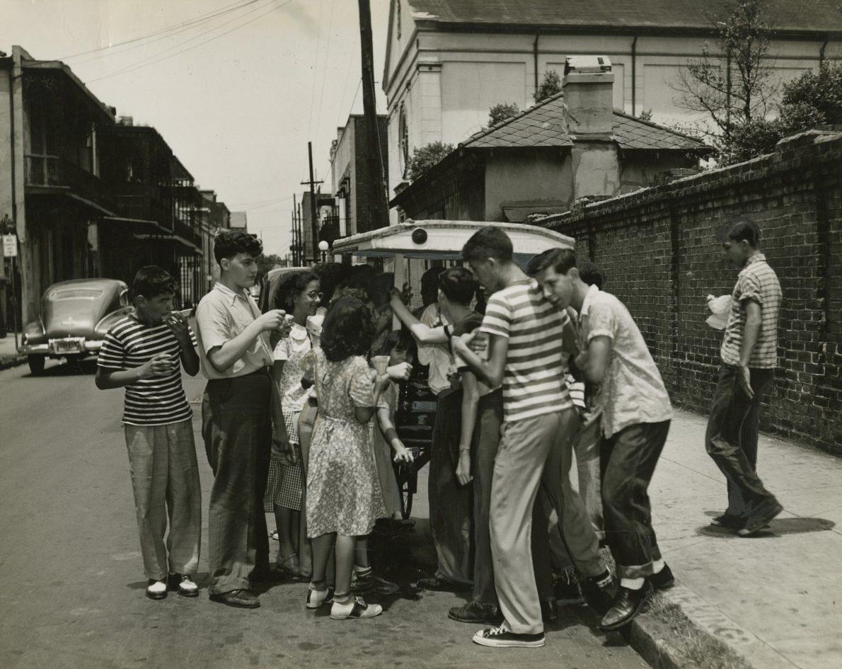 A group of people gathered around a street vendor in a lively urban setting. They are enjoying drinks, chatting, and wearing casual 1950s-style clothing. The scene is set on a sunny day with vintage cars and buildings in the background.