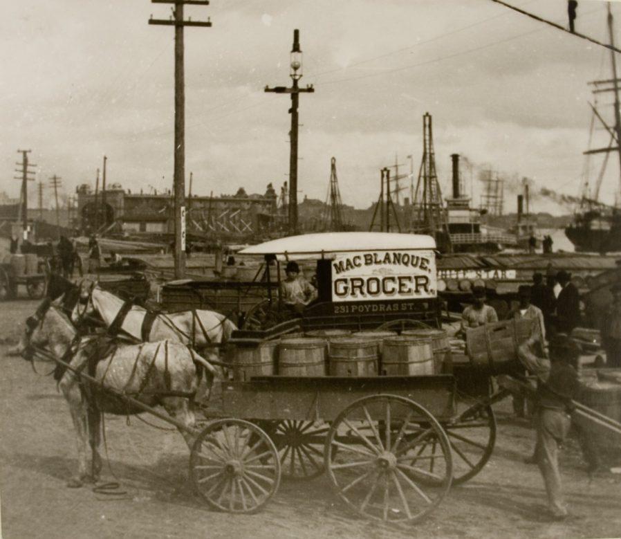 A vintage photo shows a horse-drawn wagon labeled Mac Blanque, Grocer, 231 Poydras St. on a busy dockside. People are visible handling goods, with industrial and maritime backgrounds, including ships and cranes.