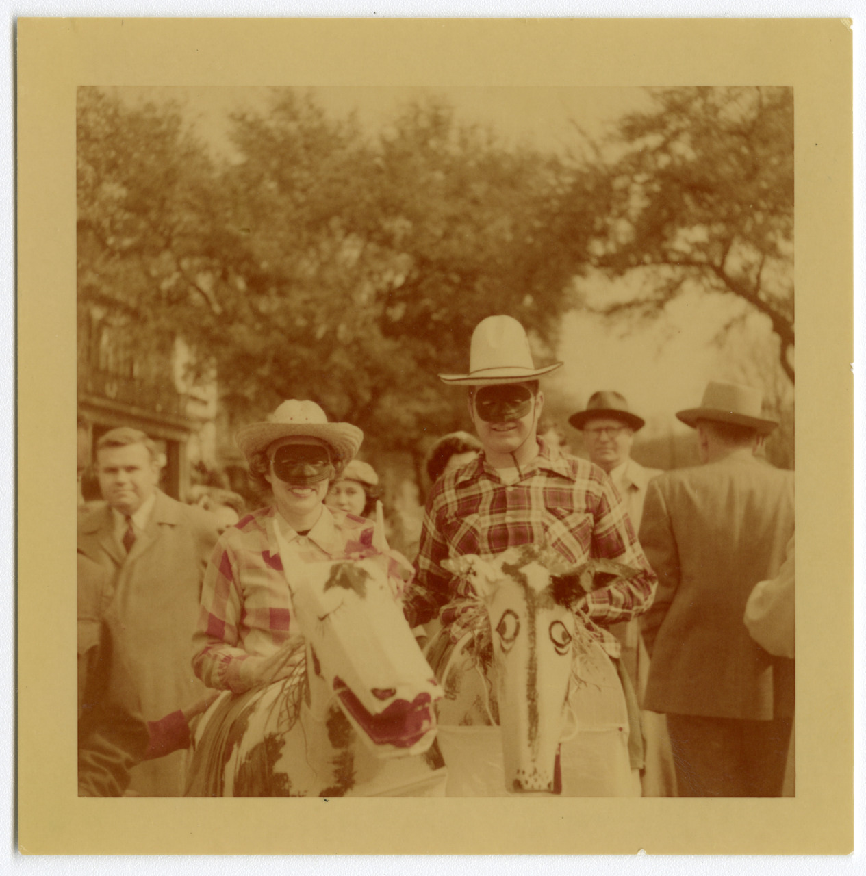 A vintage photo showing two people wearing masks and cowboy hats, standing next to each other. They are dressed in plaid shirts and riding on costume horses. A crowd of people stands around them in an outdoor setting with trees in the background.