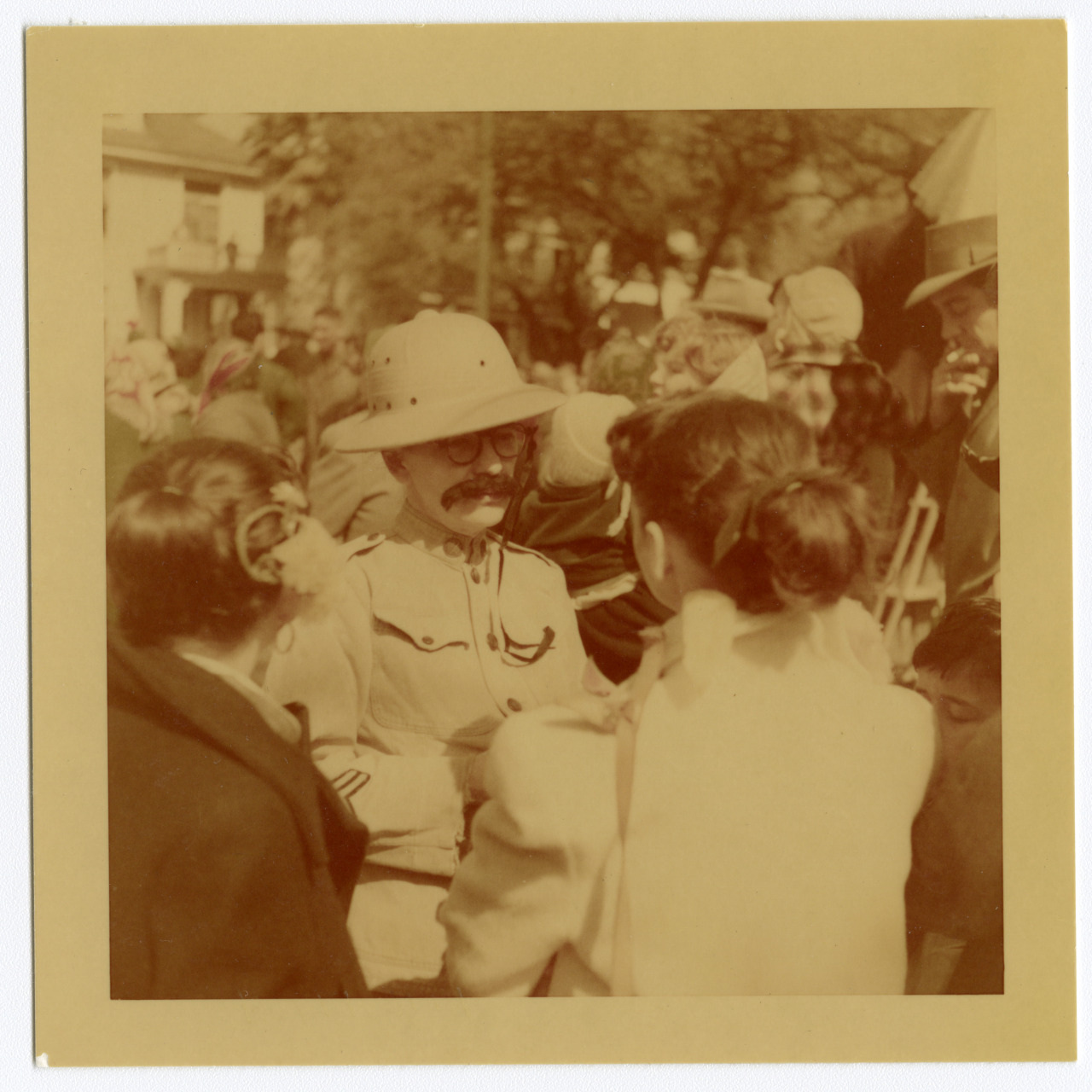 A sepia-toned photo of a man in a safari outfit and pith helmet with a fake mustache, surrounded by a crowd. Trees and buildings are visible in the background.