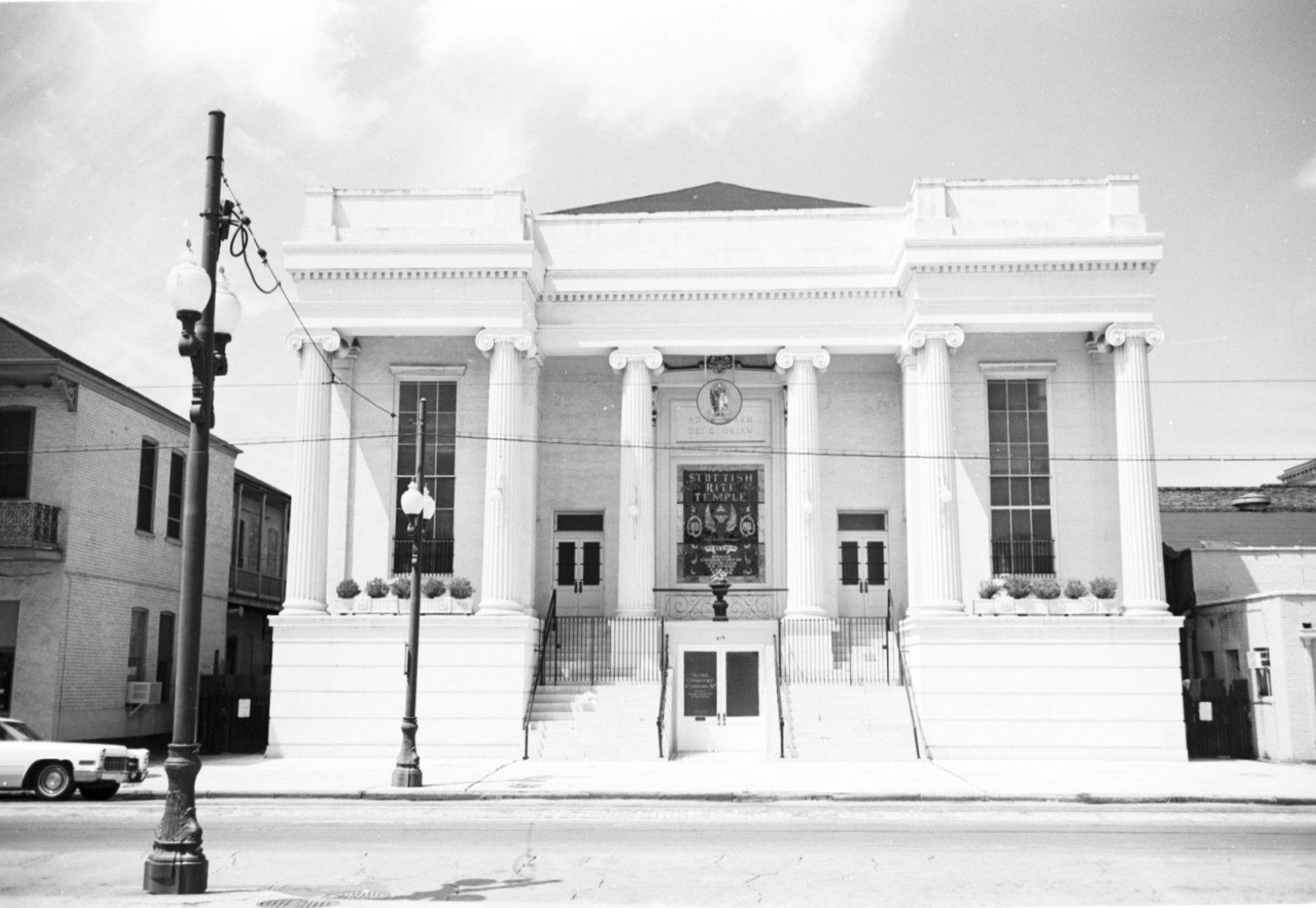 A black and white image of a historic building with tall columns, wide stairs, and ornate doors. The facade features large windows and decorative elements. A streetlight and parked car are visible in the foreground under a partly cloudy sky.