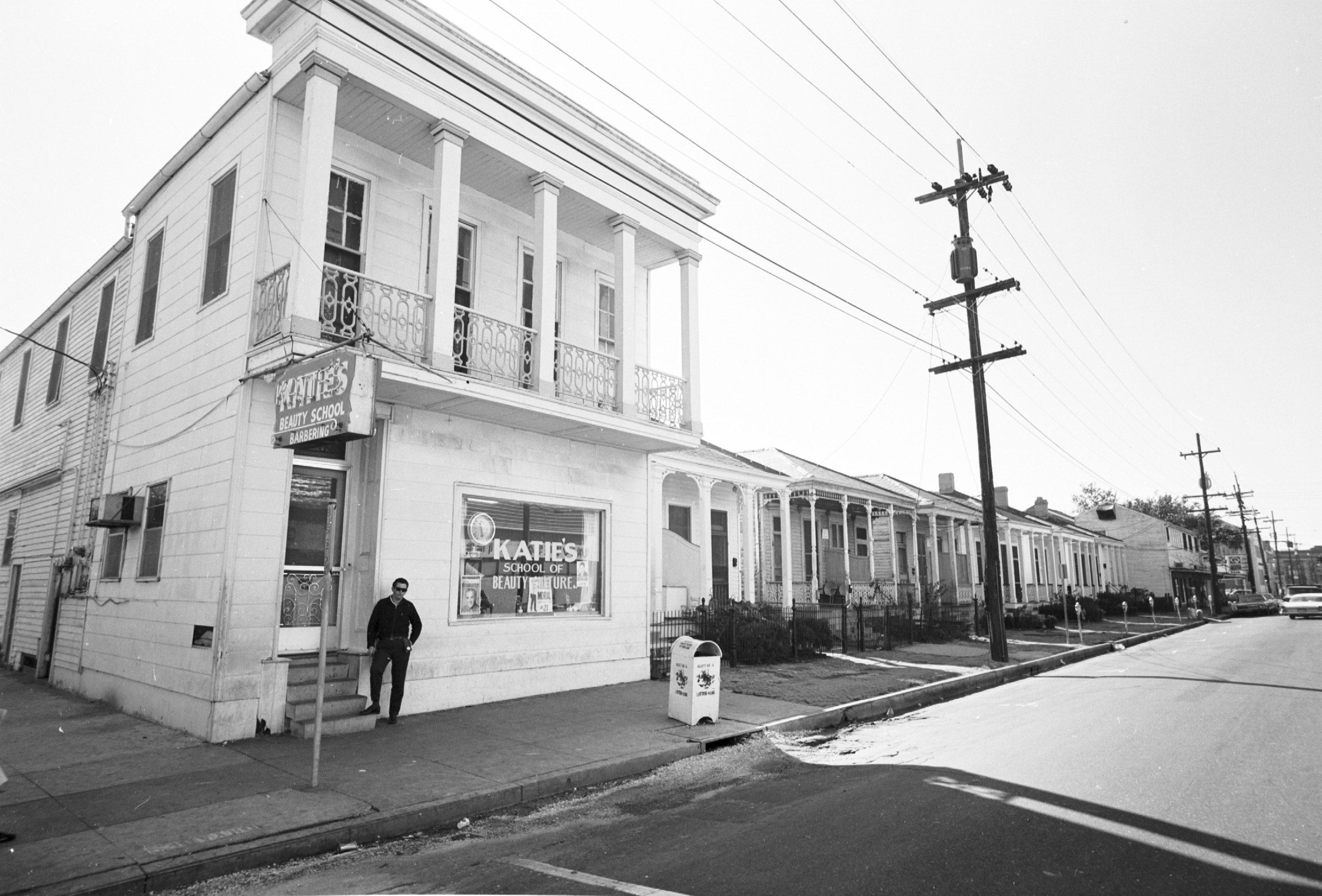 Black and white photo of a street corner with an old two-story building labeled Katies School of Hair Design. A person is walking past the entrance. The street is lined with similar buildings and power lines.