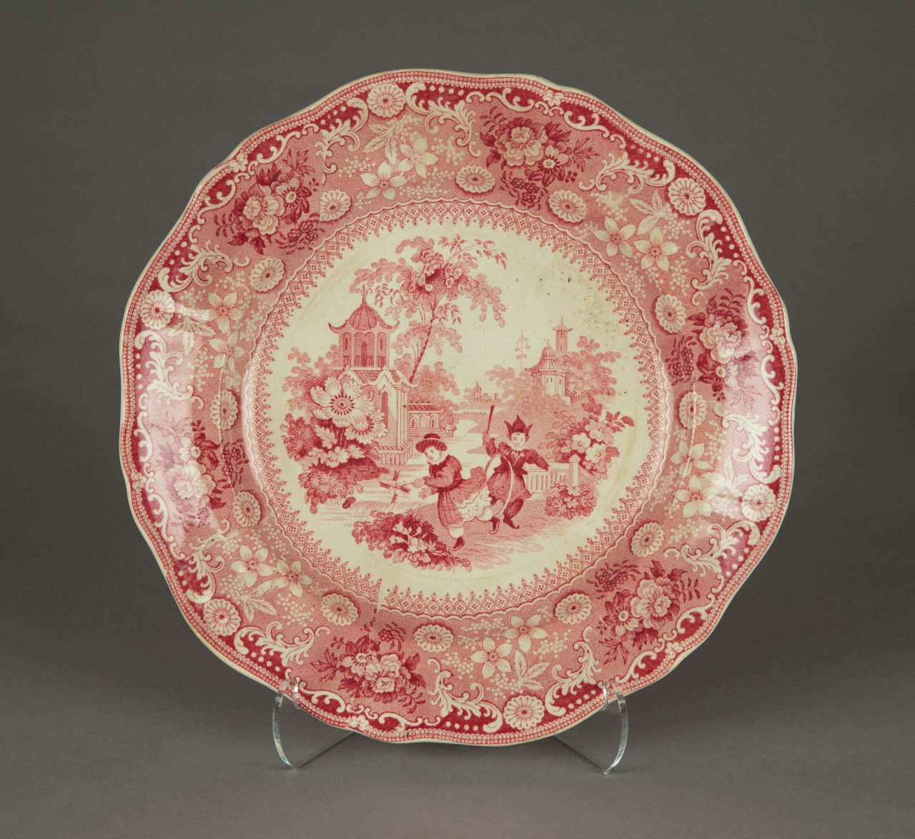 A decorative plate with a red and white pastoral scene depicting two people in a garden. The border features intricate floral patterns. The plate is displayed on a stand against a gray background.