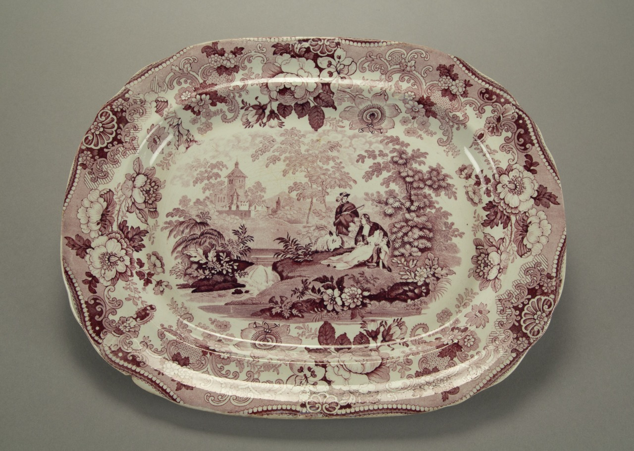 An ornate oval platter with a detailed pastoral scene in the center, featuring a man and a dog near a stream. The border has intricate floral patterns in shades of pink and maroon on a cream background.