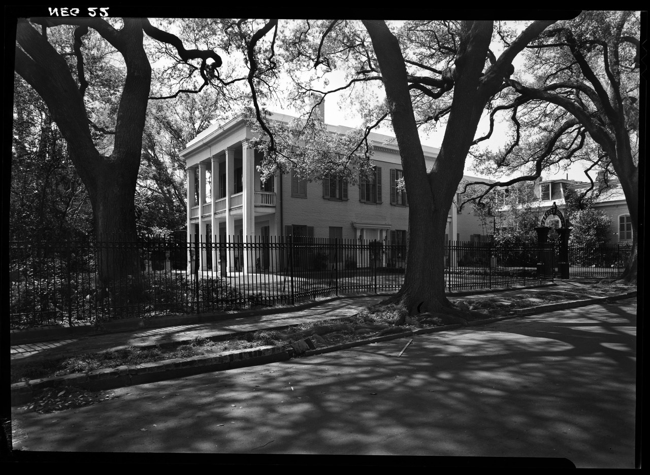 Black and white image of a large, historic house set behind an iron fence. Tall trees with spreading branches frame the view, casting shadows on the street. The house features tall columns and large windows, emphasizing its grand architectural style.