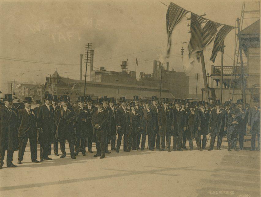 A sepia-toned photograph depicting a group of men in formal attire and top hats, standing in a line. They seem to be at a formal event, with American flags displayed prominently. A large ship and industrial buildings are visible in the background.